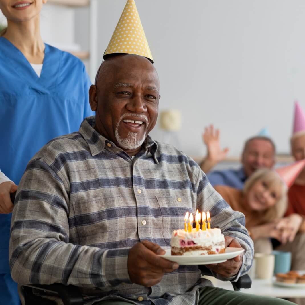 Older African American male, smiling, holding birthday cake, wearing birthday hat