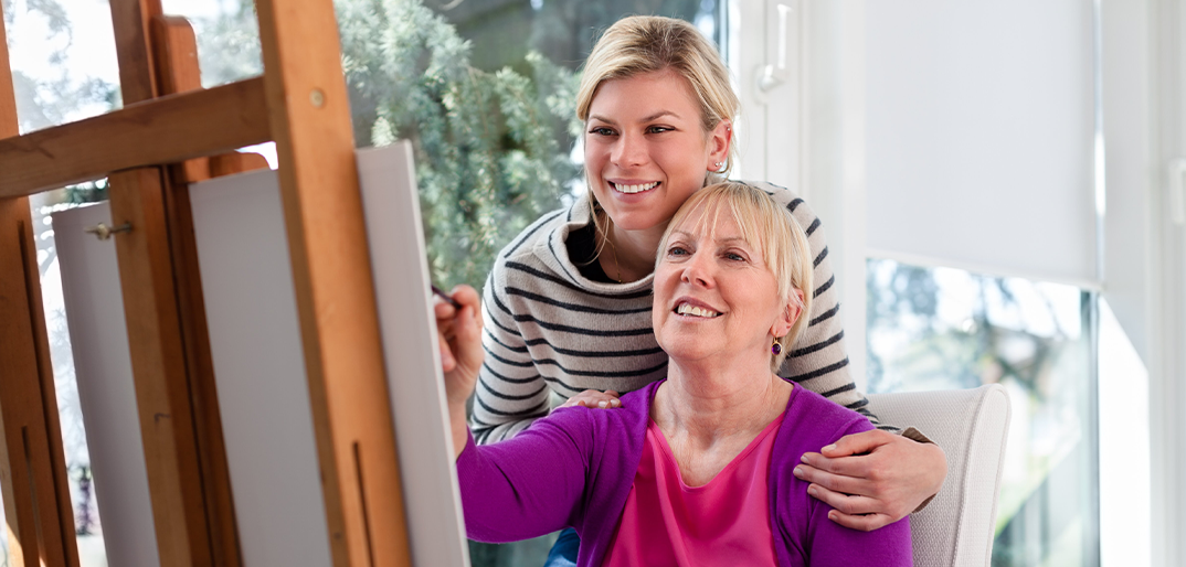 Middle aged woman standing over older woman painting, both smiling